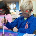 A boy in a blue sweatshirt wearing safety goggles places a magnet with tongs