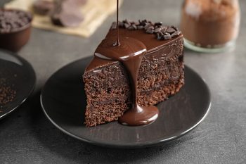 Chocolate sauce being poured onto a slice of chocolate cake, with other chocolate-based products in the background.